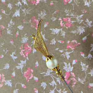 May include: A gold-toned hat pin with a pearl-like bead and a leaf-shaped design.