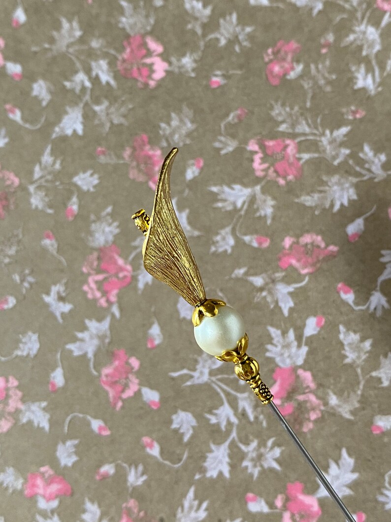 May include: A gold-toned hat pin with a pearl-like bead and a leaf-shaped design.