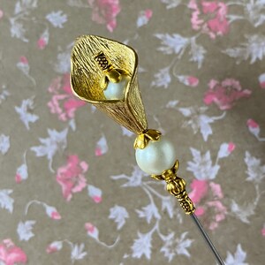 May include: A gold-toned metal flower pin with a white pearl accent. The flower is shaped like a calla lily and has a textured surface.