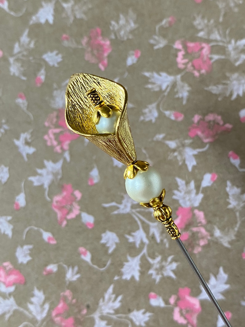 May include: A gold-toned metal flower pin with a white pearl accent. The flower is shaped like a calla lily and has a textured surface.