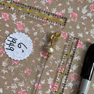 May include: A gold-colored flower pin with a pearl accent. The pin is 6 inches long and is on a brown background with a white circle that says "6" and "bthdy".