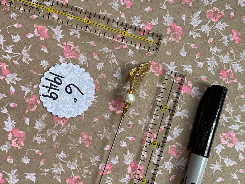 May include: A gold-colored flower pin with a pearl accent. The pin is 6 inches long and is on a brown background with a white circle that says "6" and "bthdy".