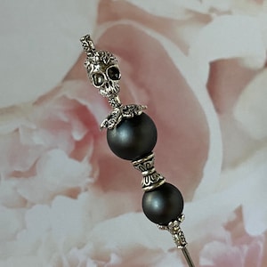 May include: A silver skull charm with ornate details sits atop two dark gray beads on a silver pin. The pin is set against a pink and white floral background.