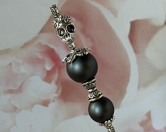 May include: A silver skull charm with ornate details sits atop two dark gray beads on a silver pin. The pin is set against a pink and white floral background.