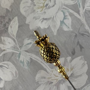 Op de afbeelding: Een gouden ananasvormige broche met een lange, dunne steel. De ananas is gedetailleerd met een getextureerd oppervlak en een kleine gouden kroon.
