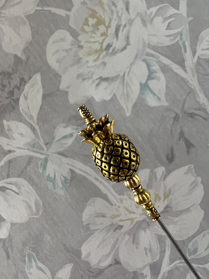 Op de afbeelding: Een gouden ananasvormige broche met een lange, dunne steel. De ananas is gedetailleerd met een getextureerd oppervlak en een kleine gouden kroon.