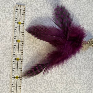 K&ouml;nnte beinhalten: Ein dekoratives Katzenspielzeug mit einem B&uuml;schel lila Federn mit schwarz-violetten Tupfen. Das Spielzeug ist an einer silberfarbenen Metallperlenkette mit dekorativen Perlen befestigt. Das Spielzeug ist etwa 10 cm lang.