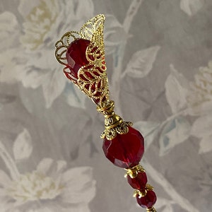 May include: A gold filigree pin with a red glass teardrop-shaped bead at the top and two smaller red glass beads below. The pin is on a white background.