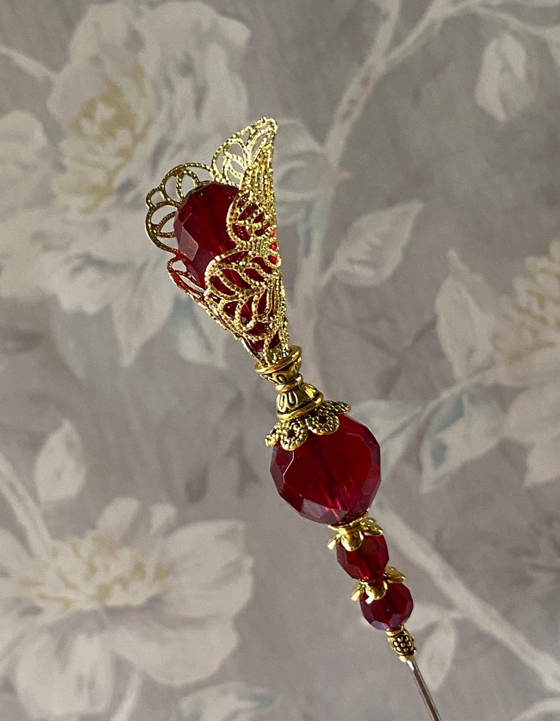 May include: A gold filigree pin with a red glass teardrop-shaped bead at the top and two smaller red glass beads below. The pin is on a white background.