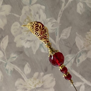 May include: A gold filigree pin with a large red faceted glass bead and two smaller red beads. The pin is on a white background with a floral pattern.