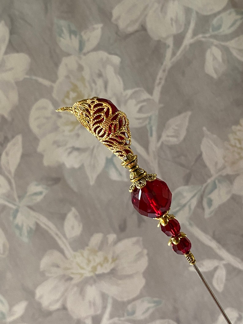 May include: A gold filigree pin with a large red faceted glass bead and two smaller red beads. The pin is on a white background with a floral pattern.