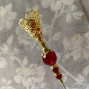 May include: A gold filigree pin with a red glass bead and a long, thin needle. The pin is designed in a floral shape.