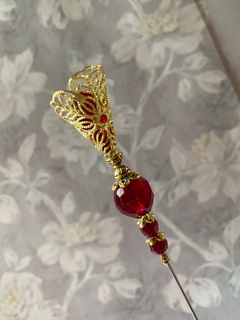 May include: A gold filigree pin with a red glass bead and a long, thin needle. The pin is designed in a floral shape.