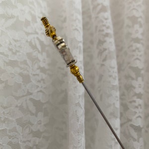 May include: A decorative hat pin with a silver shaft and gold-coloured metal detailing. The pin has an intricate design at the top and bottom, adding a touch of elegance. The background is a white lace fabric.