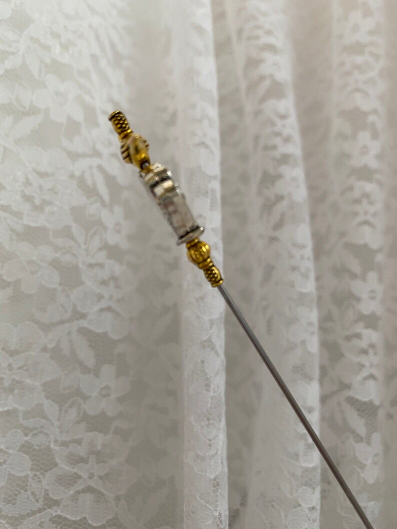 May include: A decorative hat pin with a silver shaft and gold-coloured metal detailing. The pin has an intricate design at the top and bottom, adding a touch of elegance. The background is a white lace fabric.