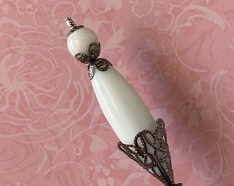 May include: A white ceramic and silver pin with a decorative design. The pin has a long, thin stem and a round, white ceramic head.