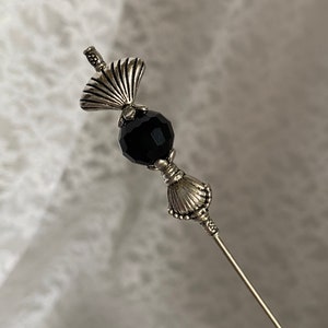 Puede incluir: Un alfiler de plata con una cuenta de cristal negro y dos abalorios decorativos en forma de concha.