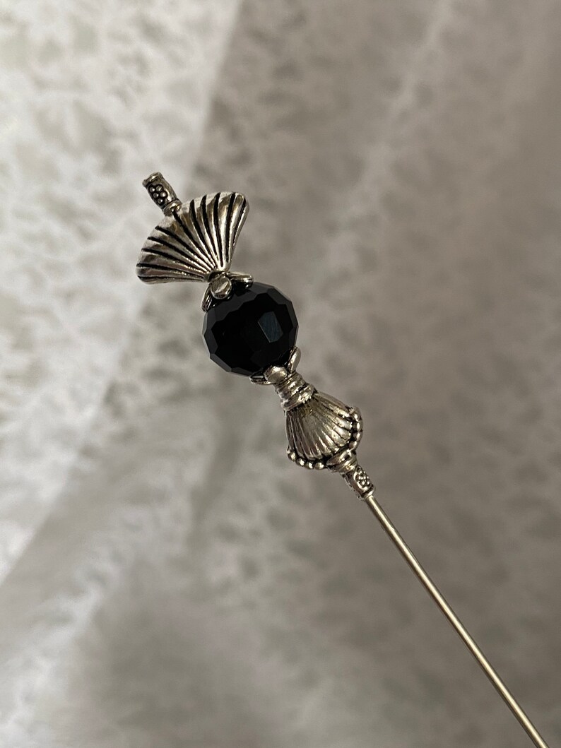 Puede incluir: Un alfiler de plata con una cuenta de cristal negro y dos abalorios decorativos en forma de concha.