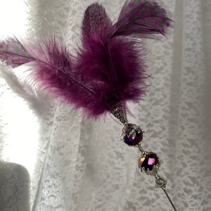 K&ouml;nnte beinhalten: Dekoratives Haaraccessoire mit Federn und einem silberfarbenen Metallstiel. Das Accessoire verf&uuml;gt &uuml;ber leuchtend violette Federn und zwei facettierte, irisierende violette Perlen. Die Metallarbeiten haben ein verziertes, vintage-inspiriertes Design.