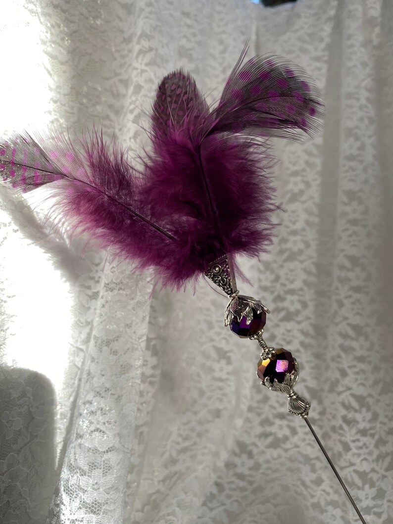 K&ouml;nnte beinhalten: Dekoratives Haaraccessoire mit Federn und einem silberfarbenen Metallstiel. Das Accessoire verf&uuml;gt &uuml;ber leuchtend violette Federn und zwei facettierte, irisierende violette Perlen. Die Metallarbeiten haben ein verziertes, vintage-inspiriertes Design.