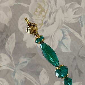 May include: A green glass pin with gold accents. The pin has a decorative top and is strung with faceted beads.