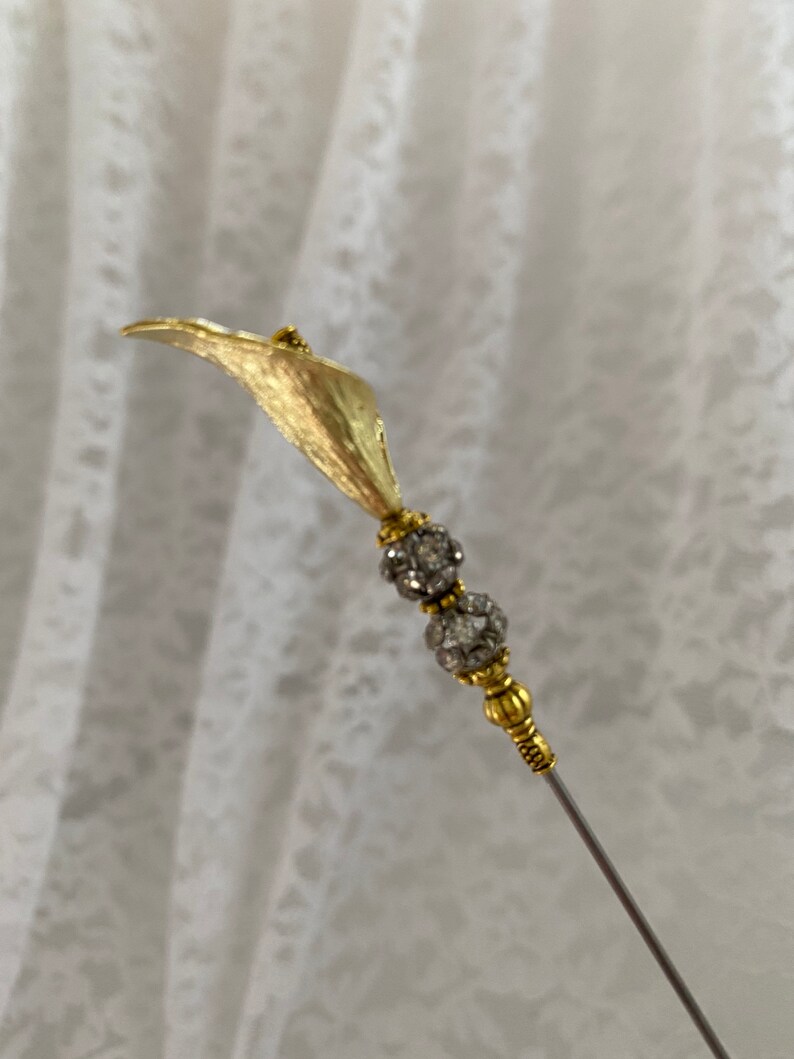 以下が含まれることがあります： 1つの花と2つの丸いクリアなラインストーンのアクセントが付いた、ゴールドトーンの金属製の花のピン