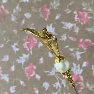 May include: A gold-toned metal pin with a white pearl bead and a stylized flower design. The pin is on a floral patterned background.