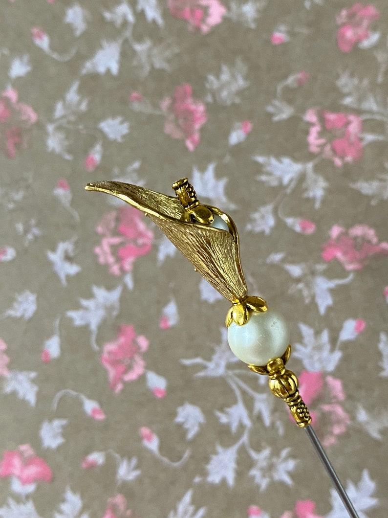 May include: A gold-toned metal pin with a white pearl bead and a stylized flower design. The pin is on a floral patterned background.