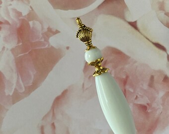 May include: A white glass pin with a gold metal top and a decorative bead. The pin is on a pink and white floral background.