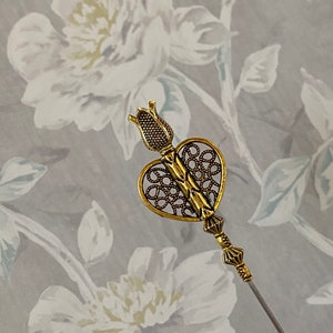 May include: A gold-toned heart-shaped pin with a decorative filigree design. The pin has a long, thin needle and a small, ornate flower-shaped top.