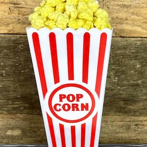 May include: A classic popcorn container filled with yellow popcorn. The container is white with red vertical stripes and a red circle with the words "POP CORN". The container is sitting on a black and white striped surface.