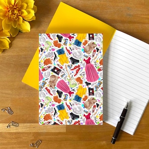 May include: A notebook with a white cover featuring a colourful pattern of various items, including clothing, shoes, and musical instruments. The notebook is open to a lined page, with a yellow envelope and a pen nearby.