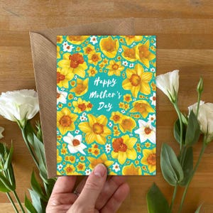 May include: A rectangular greeting card with a teal background featuring a pattern of yellow daffodils and white flowers. The text "Happy Mother's Day" is printed in white script in the center. The card is partially tucked into a brown envelope.