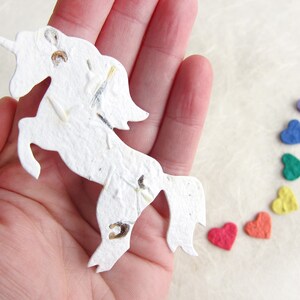 May include: A white paper unicorn with a horn and a mane made of seeds. The unicorn is standing on a white surface with a rainbow of colorful paper hearts.