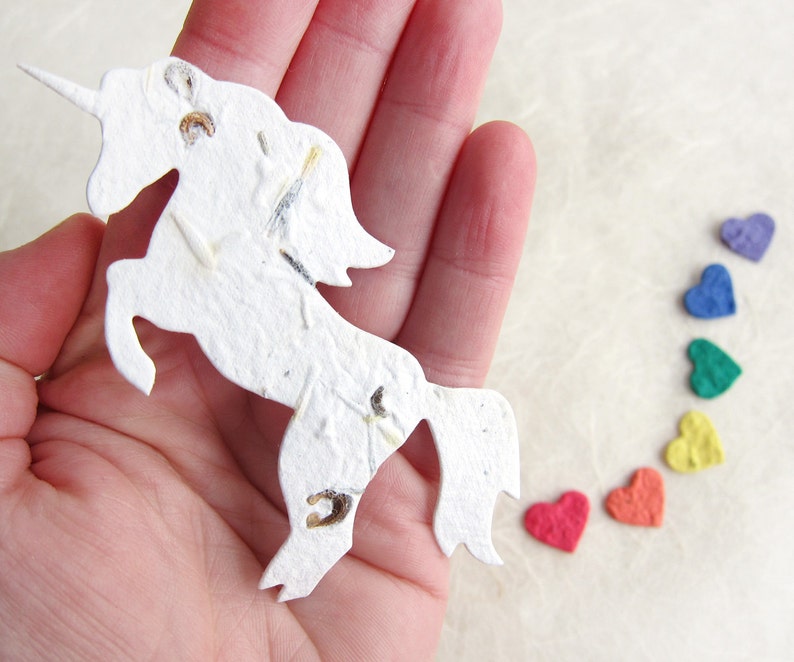 May include: A white paper unicorn with a horn and a mane made of seeds. The unicorn is standing on a white surface with a rainbow of colorful paper hearts.