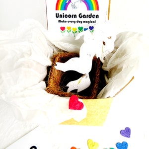 May include: A white unicorn shaped paper pot with a rainbow and the text "Unicorn Garden Make every day magical" on the box. The pot is sitting in a brown nest-like material. There are six small felt hearts in rainbow colors on a white surface.