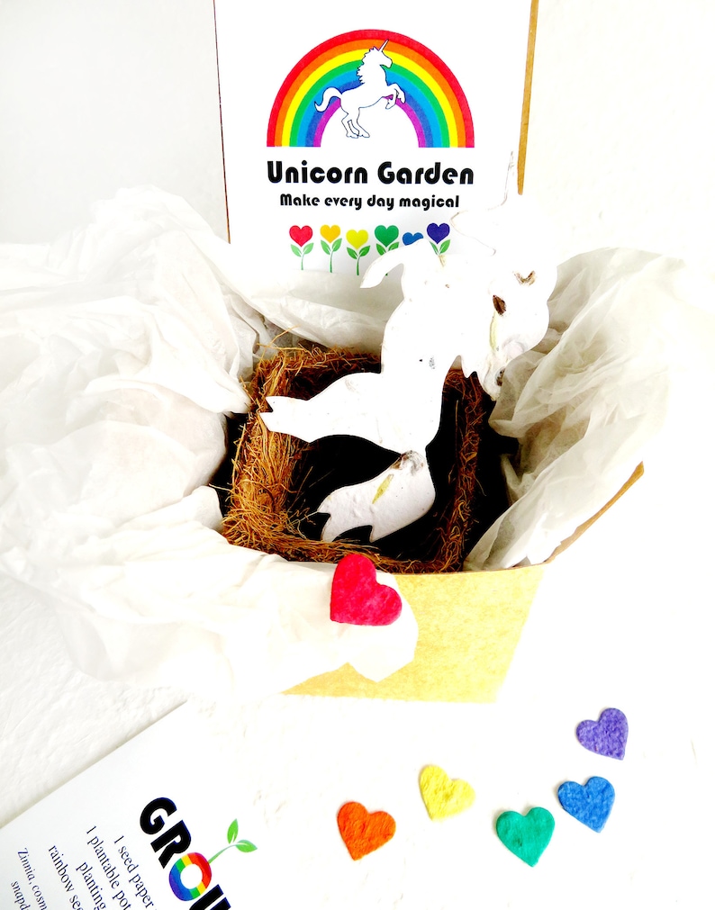 May include: A white unicorn shaped paper pot with a rainbow and the text "Unicorn Garden Make every day magical" on the box. The pot is sitting in a brown nest-like material. There are six small felt hearts in rainbow colors on a white surface.