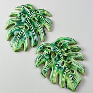 May include: Two ceramic wall decorations shaped like monstera leaves. Each leaf features a glossy, variegated glaze in shades of emerald, lime, and forest green with deep indentations and natural-looking perforations throughout the foliage.