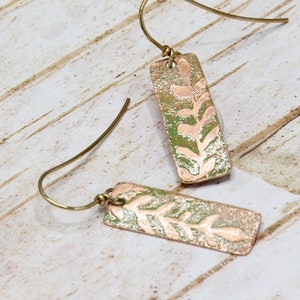 May include: A pair of copper earrings with a green and pink floral design. The earrings are rectangular in shape and have a simple wire hook.