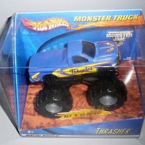Może przedstawiać: Niebiesko-ż&oacute;łty monster truck Hot Wheels Monster Jam Thrasher w przezroczystym plastikowym pudełku. Ciężar&oacute;wka ma czarne koła, a pudełko ma napis "Monster Jam" i "Thrasher".