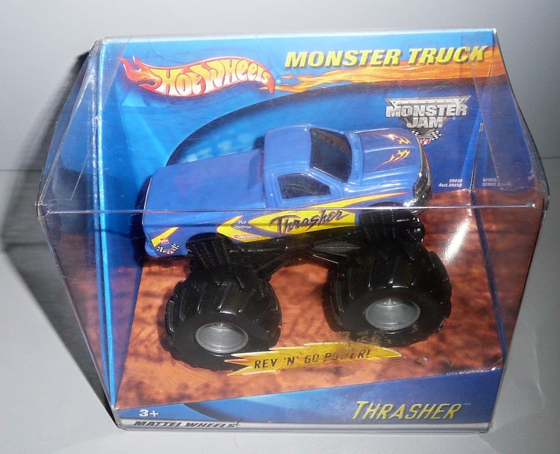 Może przedstawiać: Niebiesko-ż&oacute;łty monster truck Hot Wheels Monster Jam Thrasher w przezroczystym plastikowym pudełku. Ciężar&oacute;wka ma czarne koła, a pudełko ma napis "Monster Jam" i "Thrasher".