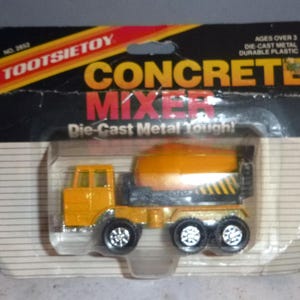 May include: A toy concrete mixer truck in its original packaging. The truck is yellow and orange with black wheels. The packaging reads "TOOTSIETOY CONCRETE MIXER Die-Cast Metal Tough!" and "AGES OVER 3".