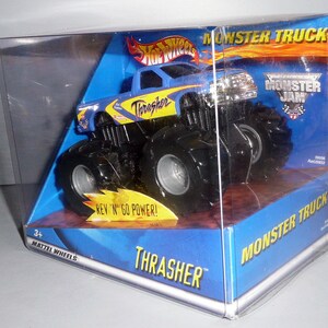 Może przedstawiać: Niebiesko-ż&oacute;łty Monster Jam, zabawkowy samoch&oacute;d ciężarowy z napisem "Thrasher" na boku. Samoch&oacute;d znajduje się w przezroczystej plastikowej gablocie z niebiesko-ż&oacute;łtym tłem. Na pudełku widnieje napis "Monster Truck" i "Rev 'N' Go Power!"
