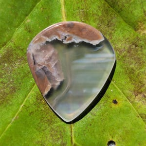 Puede incluir: Una piedra preciosa pulida en forma de lágrima, con una mezcla de colores. La piedra presenta áreas marrones, grises y blancas translúcidas, con un acabado brillante. La piedra preciosa está sobre una hoja verde.