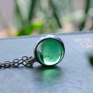 May include: A silver necklace with a large, round, transparent green pendant. The pendant is set in a hammered metal bezel and hangs from a delicate chain. The background is blurred, suggesting an outdoor setting.