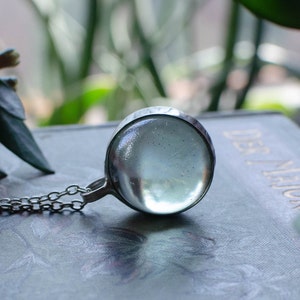 May include: A silver pendant necklace with a clear, round glass orb. The orb is set in a metal bezel and suspended from a fine chain. The background is blurred, suggesting a natural setting.