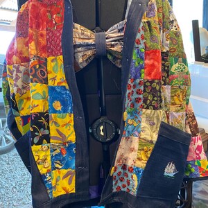 May include: A patchwork jacket with a denim collar, cuffs, and pockets. The jacket features a variety of colourful fabric squares with floral, insect, and whimsical designs. A bow tie is attached to the mannequin. The pocket has an embroidered sailboat.