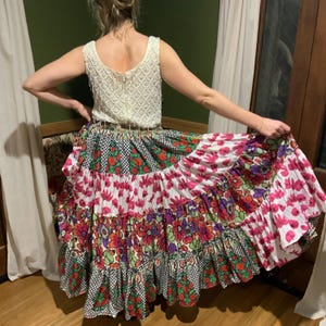 May include: A cream-coloured, sleeveless top with a textured, scalloped design is paired with a long, tiered skirt. The skirt features a patchwork of floral and geometric patterns in shades of pink, red, green, and purple.