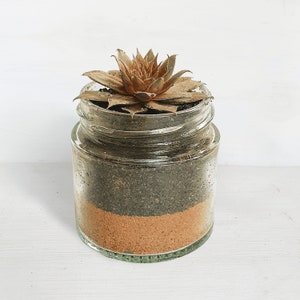 May include: A clear glass jar with a brown and tan layered soil mixture and a dried succulent plant with brown leaves.
