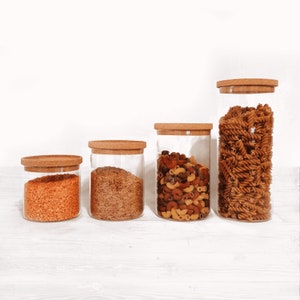 May include: Four glass jars with cork lids, filled with different dry goods. The jars are lined up in a row from smallest to largest. The smallest jar is filled with red lentils, the next jar is filled with brown rice, the next jar is filled with a mix of pasta shapes, and the largest jar is filled with brown curly pasta.